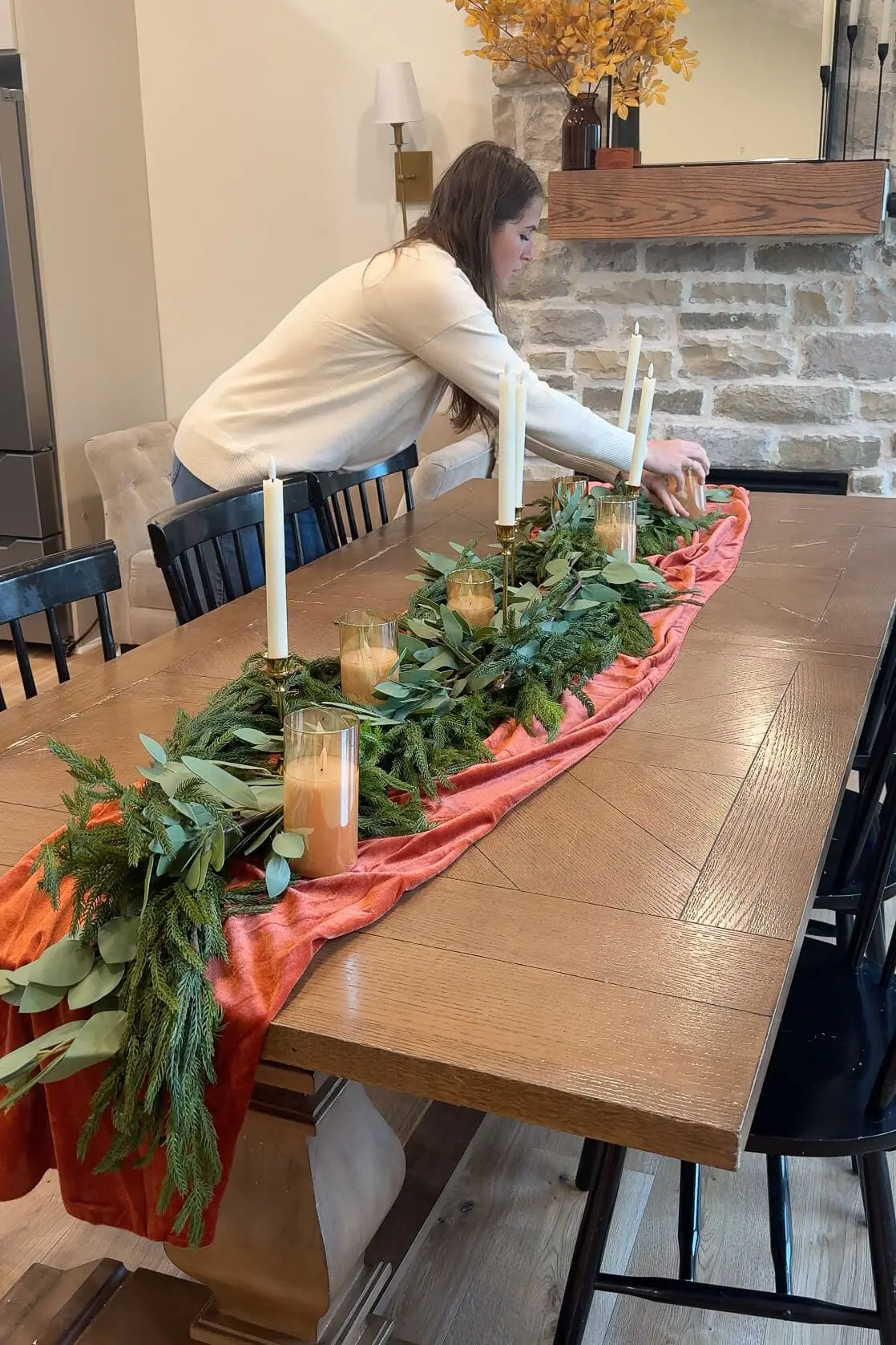 Woman adding candles in glass holders and tall brass candlesticks to a festive tablescape decorated with greenery on an orange velvet runner.
